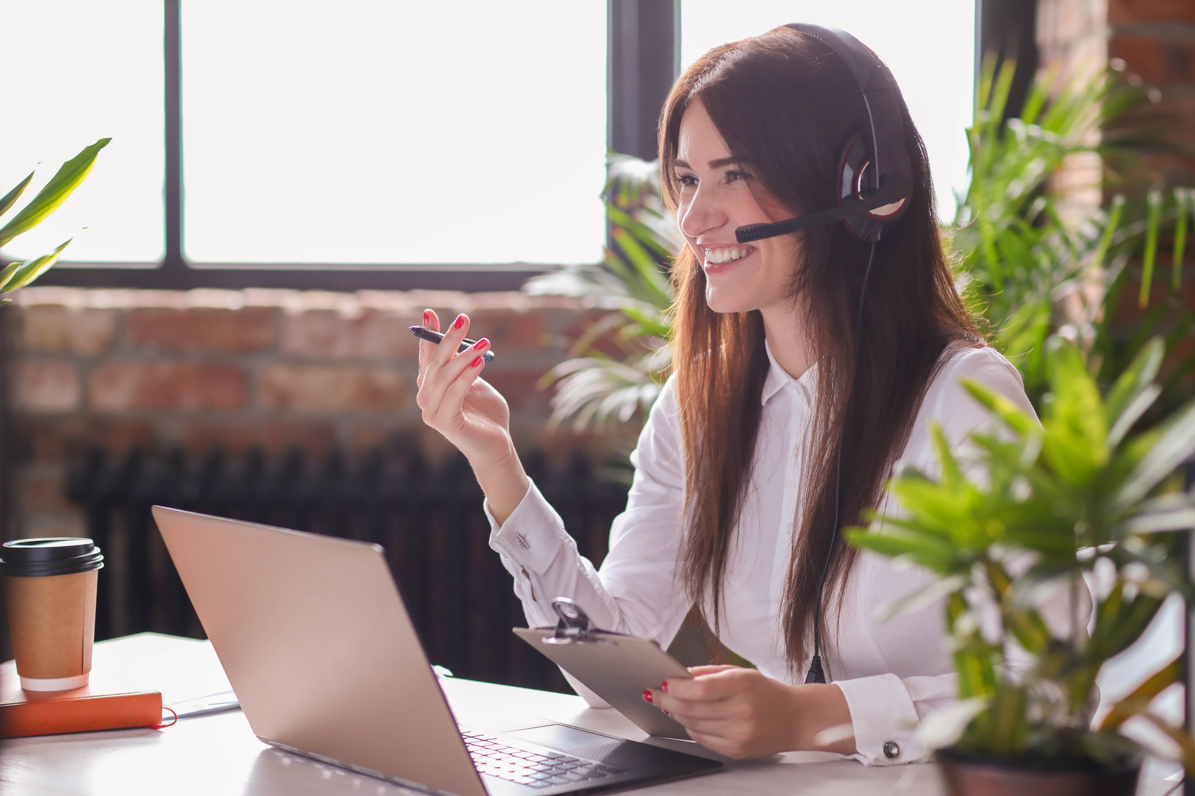 Imagem de uma mulher sorrindo, usando fones de ouvido com microfone, enquanto trabalha em um laptop. Ela segura um bloco de notas e uma caneta, parecendo estar em uma videochamada ou atendimento ao cliente. A cena está ambientada em um escritório iluminado, com plantas ao fundo, criando um ambiente acolhedor e produtivo.